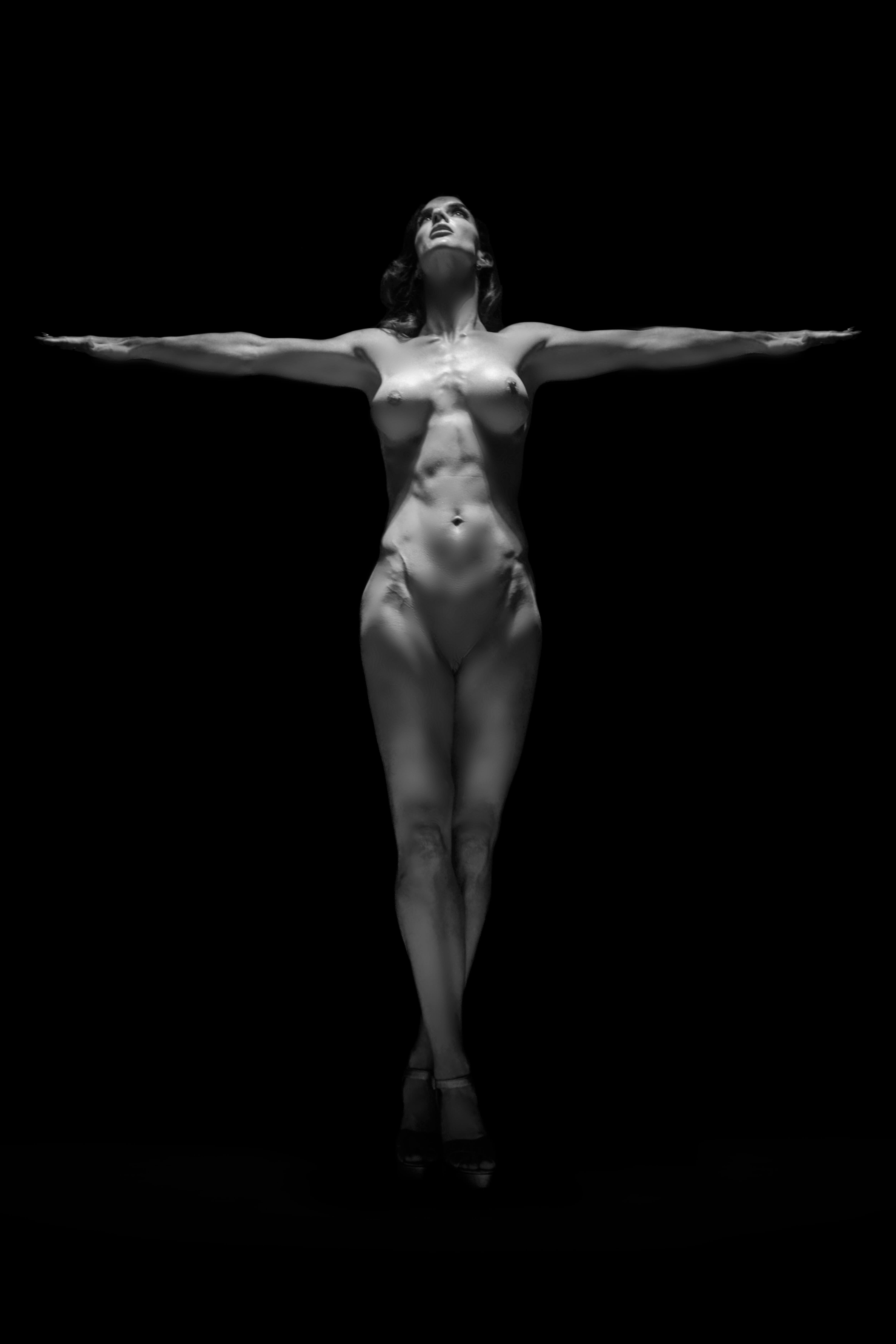 Elevation — nude female figure with arms outstretched horizontally and face lifted upward toward the light, emerging from total darkness, fine art photograph by Alex Manfredini
