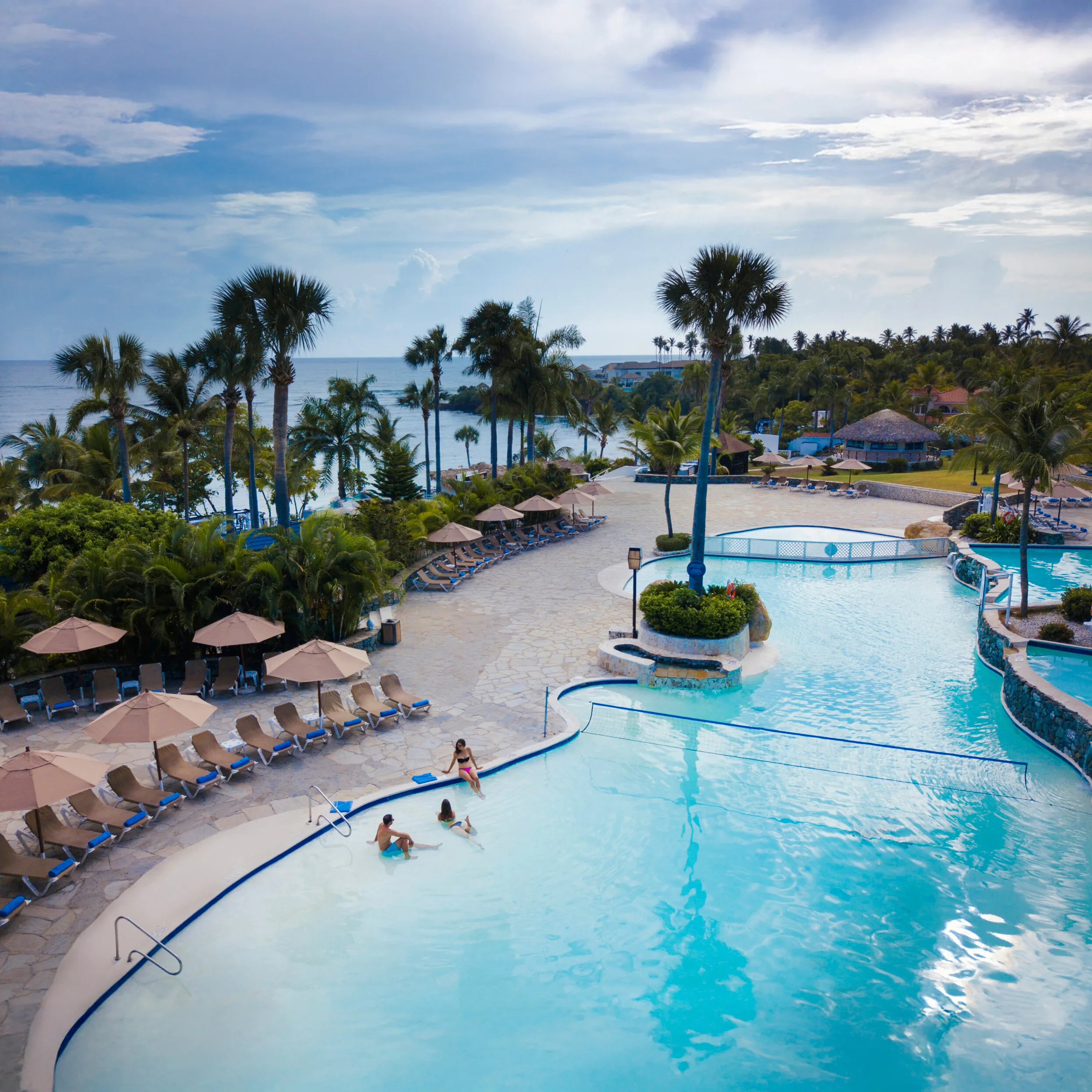 Tropical beach resort with multiple pools, palm trees, and ocean view under a partly cloudy sky, with people relaxing by and in the water.