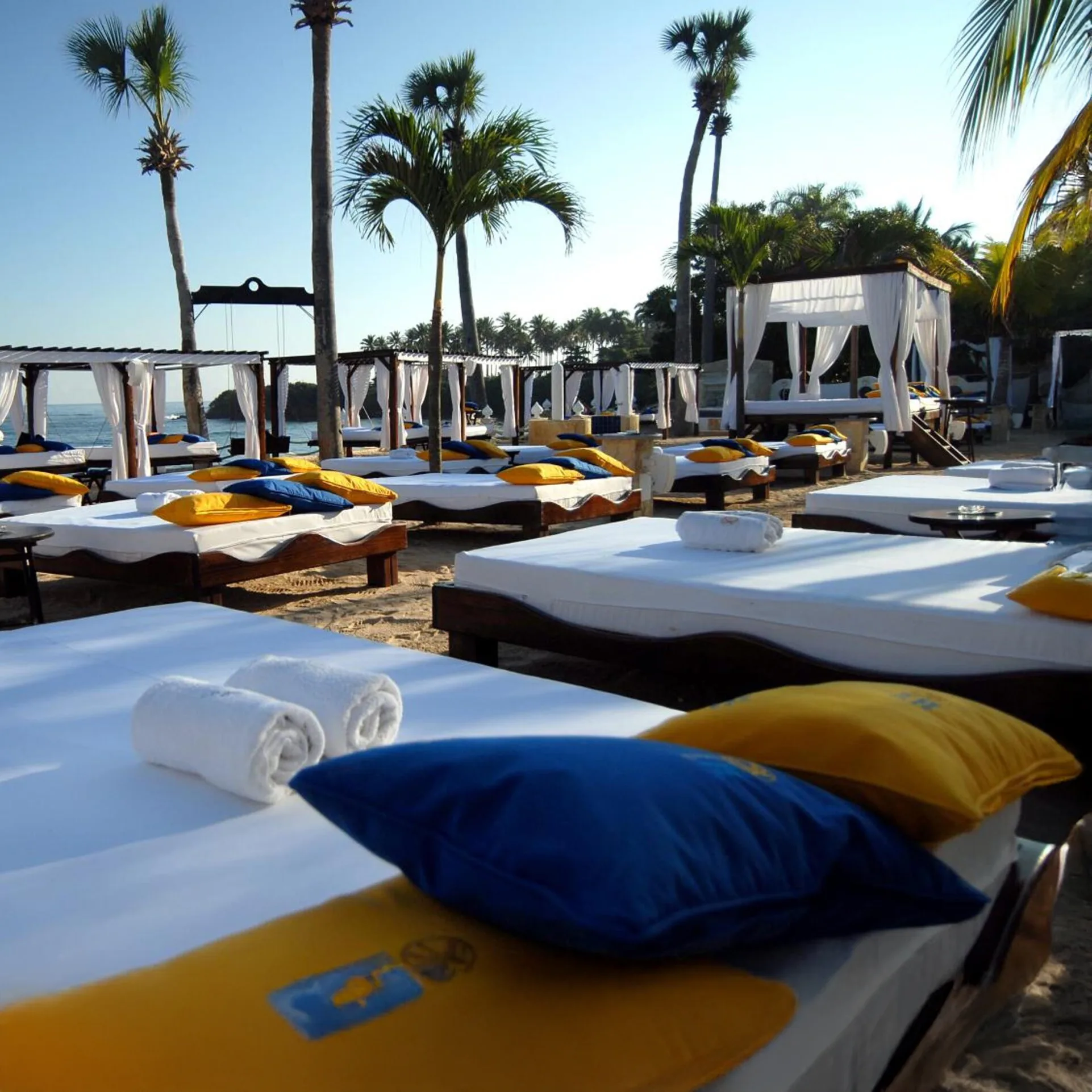 Row of luxurious white beach cabanas with blue and yellow pillows on a sandy beach under palm trees at Lifestyle Tropical Beach Resort & Spa, with the ocean and distant land visible under a clear blue sky.