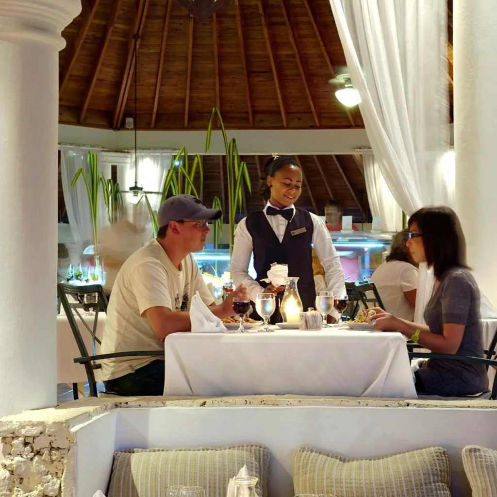 A waitress in a black vest and white shirt serves drinks to a man and woman dining at a table in a resort restaurant with a thatched roof and flowing white curtains.